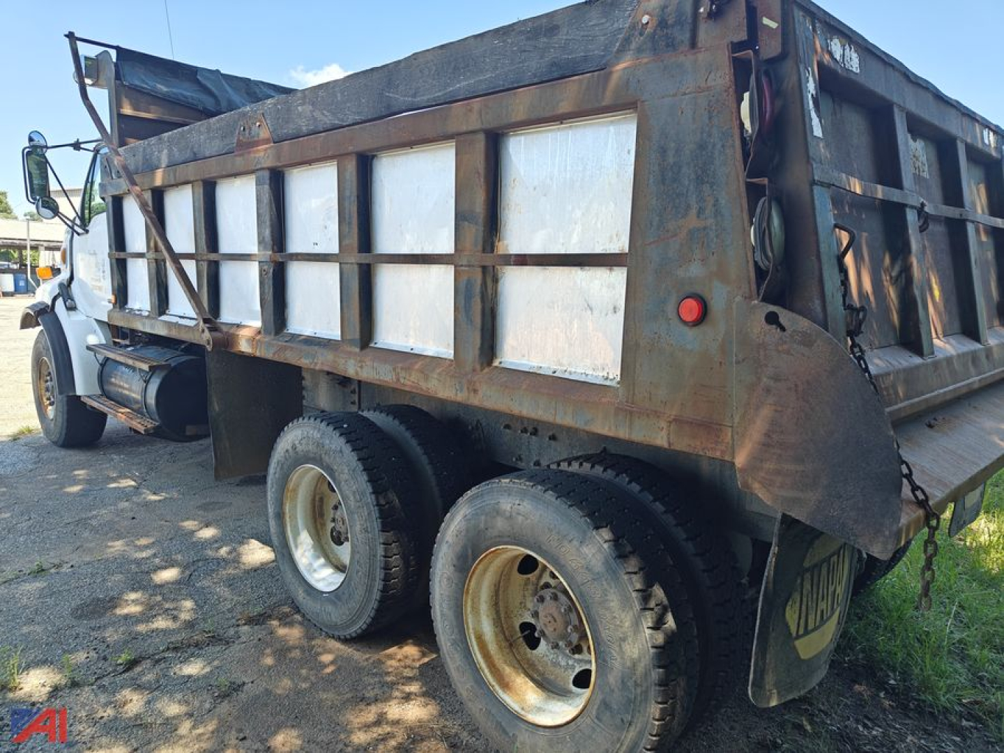 Dump truck bed side with metal side panels and dual rear wheels, rusted steel frame, rear lights, and hydraulic dump mechanism visible.