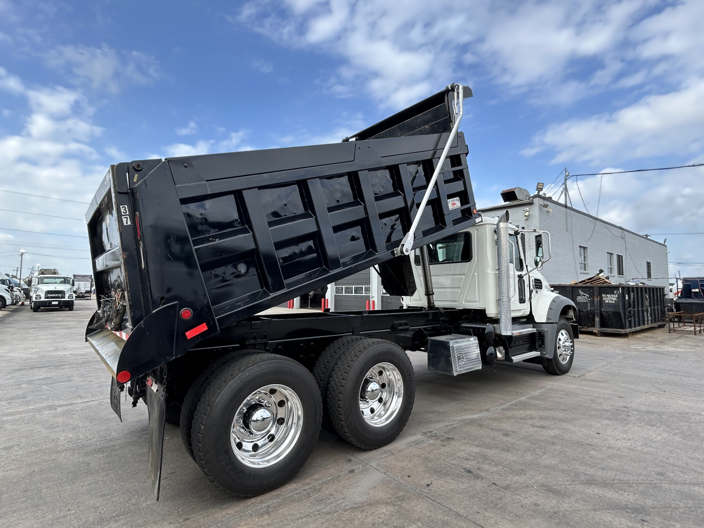 Dump truck in service showing raised black dump bed and chrome wheels, Mack GR64F with dual rear axles in a lot.