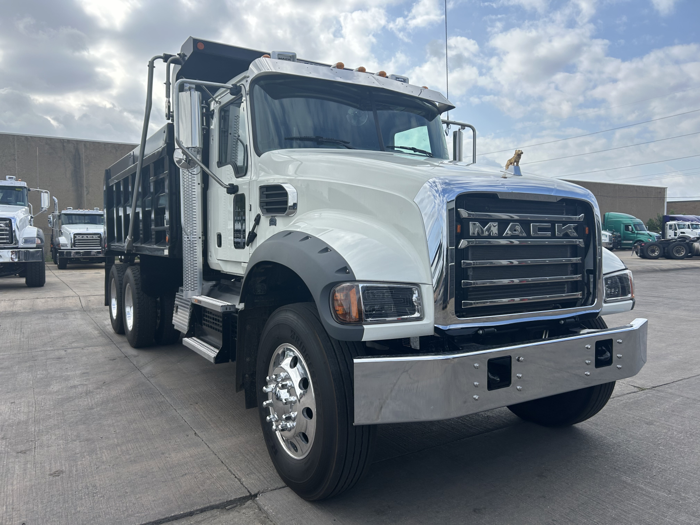 Mack GR64F dump truck in a lot, front-left view, chrome bumper, large grille with MACK letters, dual rear wheels, white cab with black fenders, cargo bed visible.