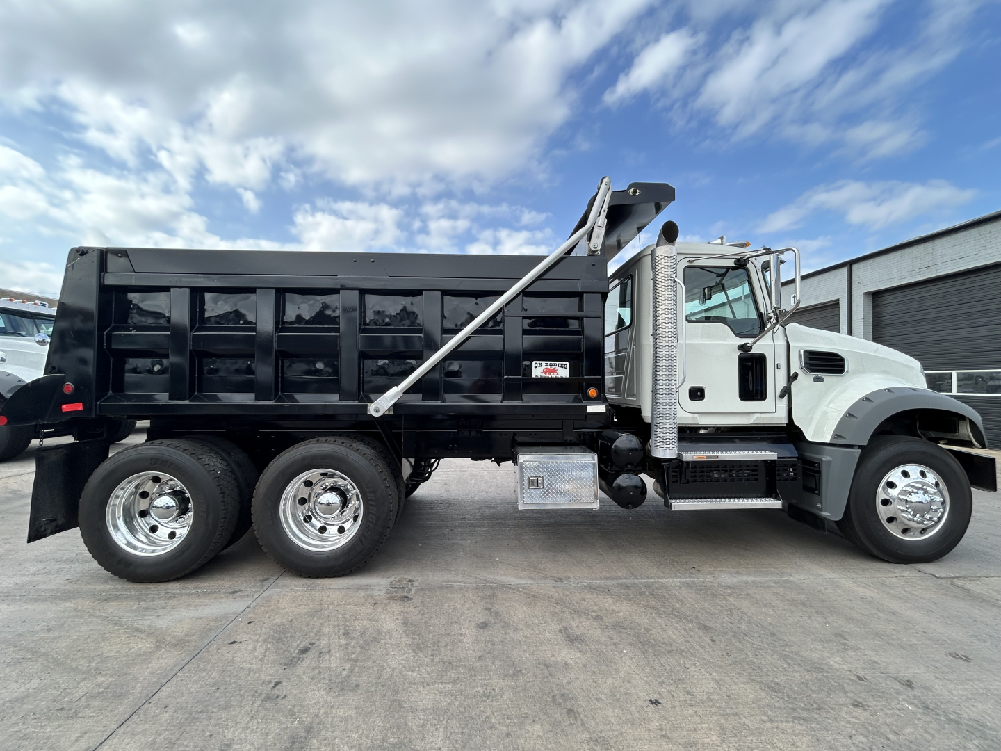 Dump truck side view showing Mack GR64F with black dump bed, chrome exhaust, and dual rear axles.