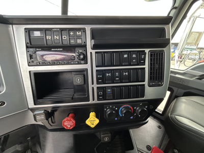 Mack truck cab dashboard showing gear selector markings R N D P plus electrical switches and climate controls; yellow and red warning knobs and a small storage compartment.