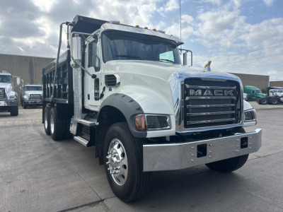 Mack GR64F dump truck in a lot, front-left view, chrome bumper, large grille with MACK letters, dual rear wheels, white cab with black fenders, cargo bed visible.