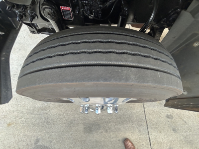 Front view of a Mack GR64F truck with a large front tire and dual wheel studs visible. Tire tread shows wear bands. Underneath are suspension components and part of the chassis.