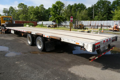 Flatbed trailer with dual rear axles and metal decking; side rails and orange reflectors; visible wheel wells and rear ramp; no legible signage or labels readable.