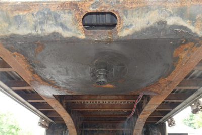 Trailer undercarriage showing rusted deck, central circular plate, and hitch mounting area with I-beam frame underneath.
