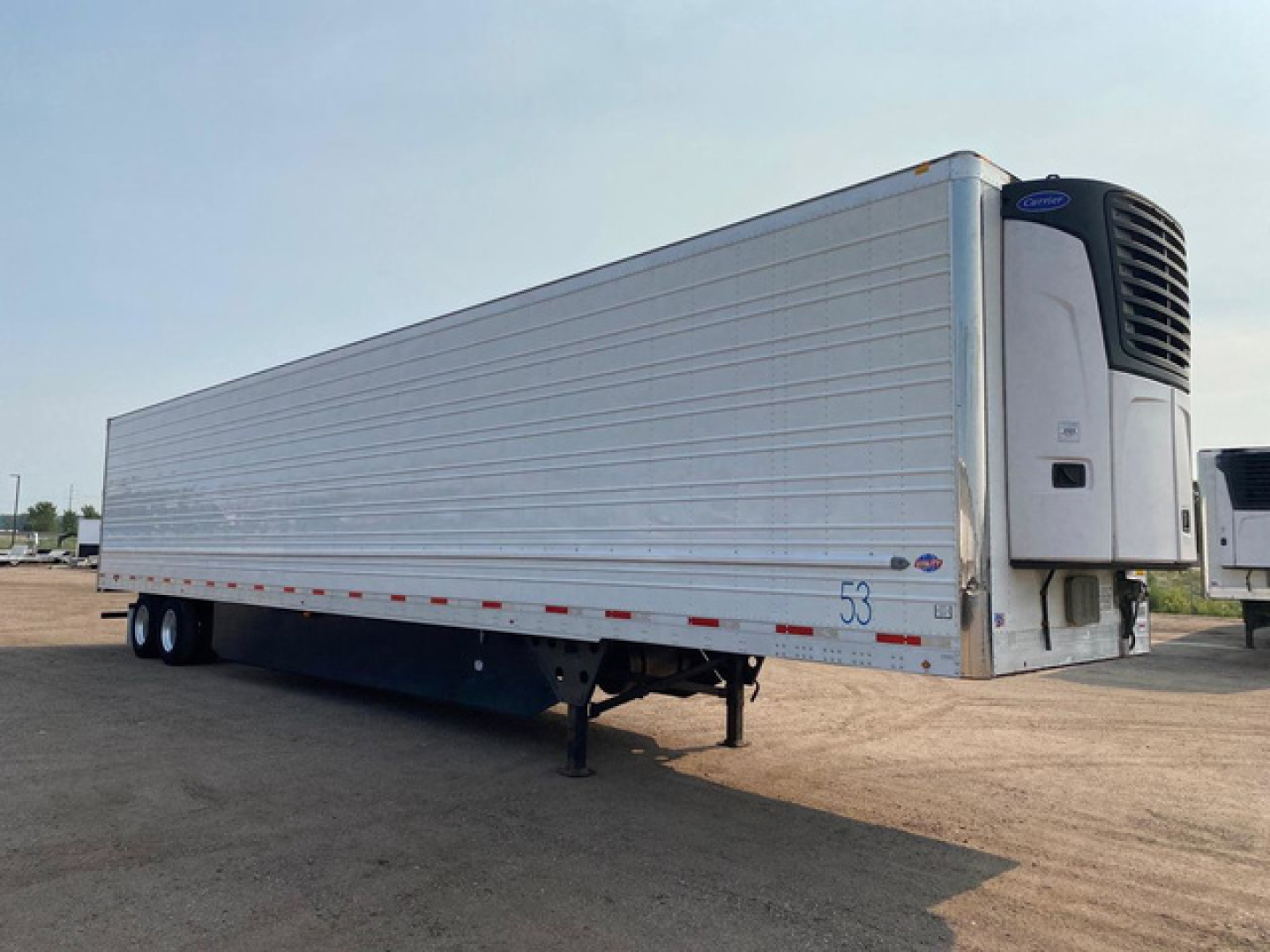 Trailer side view showing refrigerated unit at front with black cooling fan housing, aluminum siding, and undercarriage. Visible text includes '53' on door area and small decals near front.