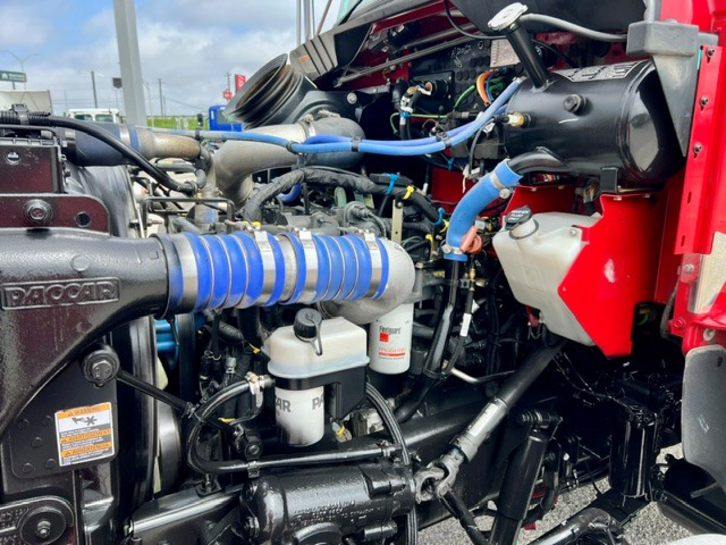 Engine bay of a Kenworth truck showing blue hoses, filters, and various engine components.
