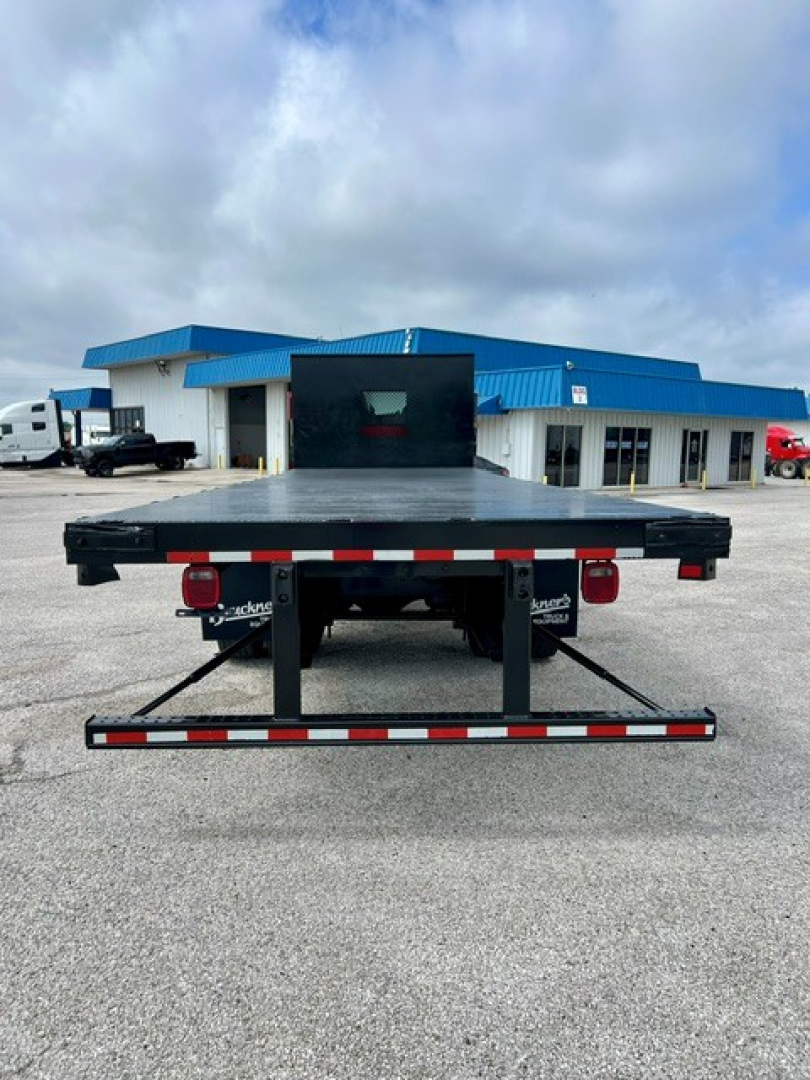 Flatbed truck rear view showing a black steel flatbed with rear bumper, taillights, and undercarriage. Kennworth/flatbed equipment visible with loading ramp and reflective tape.