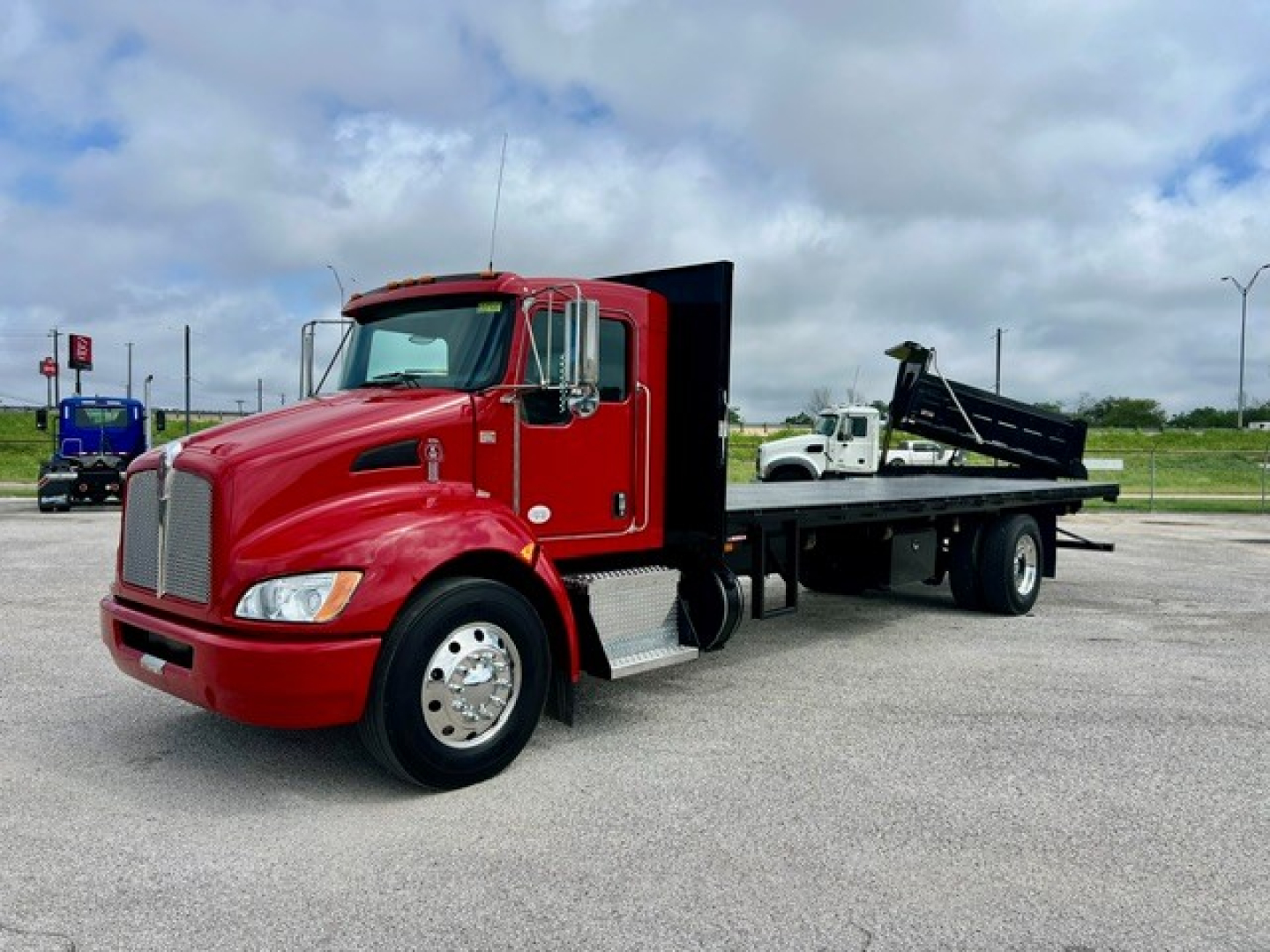 Kenworth flatbed tow truck in red with long wheelbase, cab-over style; front grille with chrome, large side mirrors, and flatbed with hydraulic dump bed.