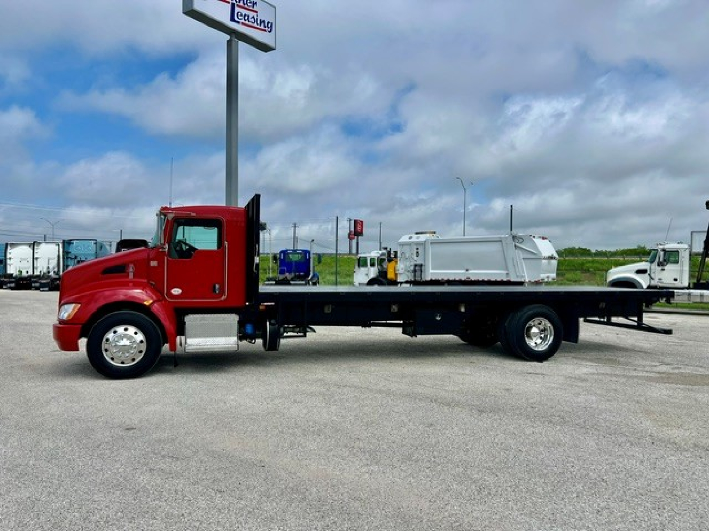 Kenworth T270 flatbed truck side view showing red cab and long black flatbed with equipment load near a dealership lot.