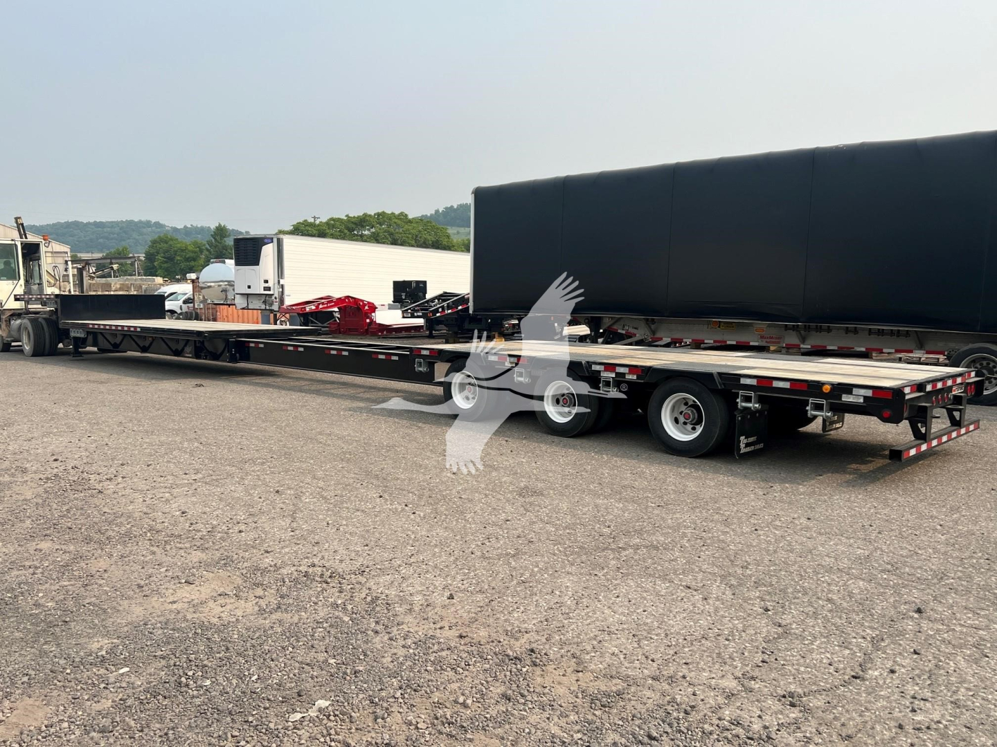 Long steel drop extendable trailer in a yard with a large black tarp cover and other equipment in background.