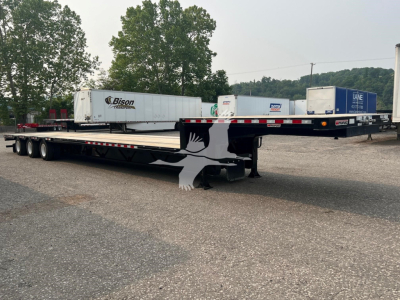 Trailer yard view of a 2023 Manac steel drop extendable trailer with long flatbed and dual rear axles, few parked trailers in background.