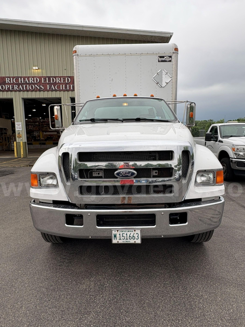 Ford F-650 front view in service lot showing chrome grille, large bumper, and box truck behind. License plate visible but unreadable.