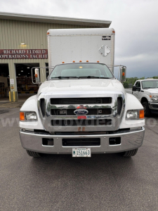 Ford F-650 front view in service lot showing chrome grille, large bumper, and box truck behind. License plate visible but unreadable.