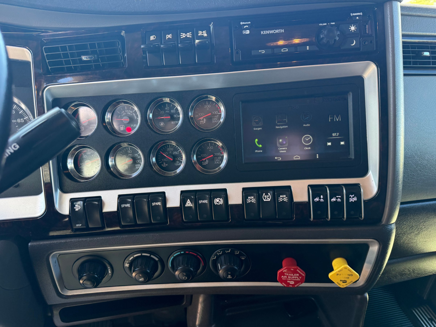 Kenworth W900L dash showing multiple analog gauges, infotainment screen, and control switches; dashboard chrome trim with red and yellow knobs.