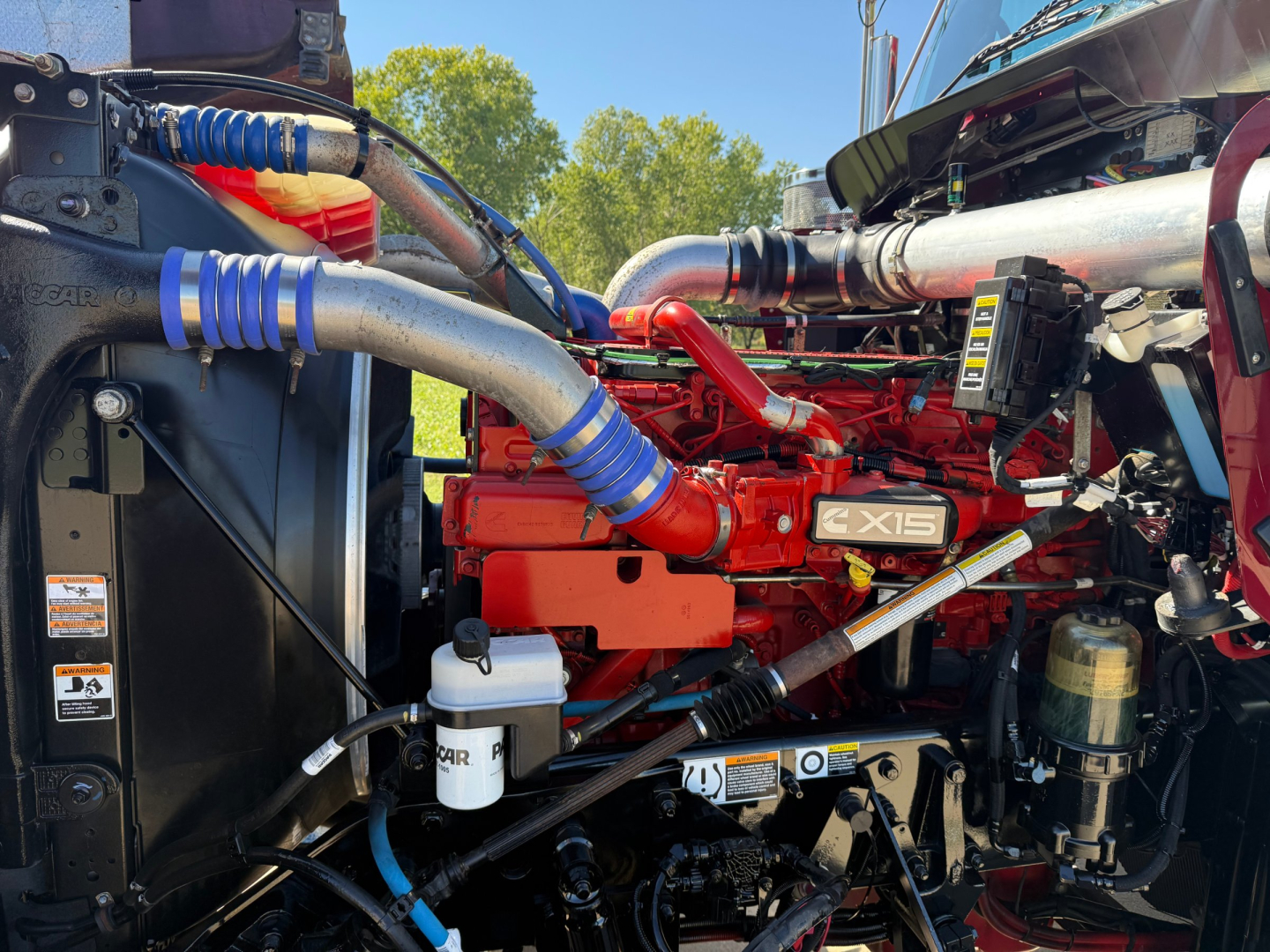 Kenworth W900L engine bay with red GLS exhaust, blue hose clamps, and multiple hoses; visible components include coolant reservoir, oil fill, and engine block