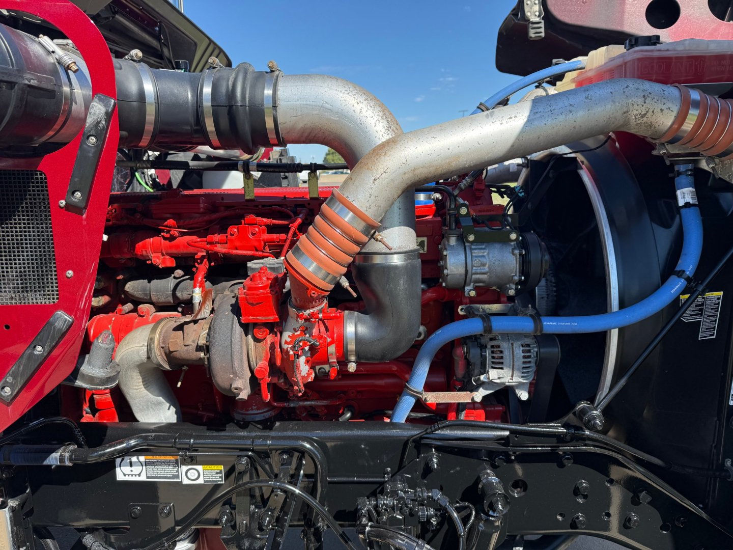 Kenworth W900L engine bay showing red engine components, large silver intake pipes, and blue hoses in view.