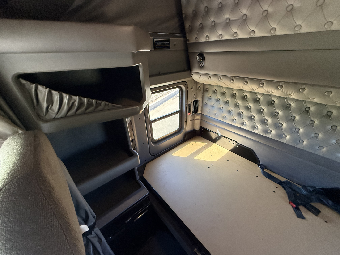 Kenworth W900L sleeper cab interior showing bunk area with quilted padded wall, small window, and overhead storage shelf.