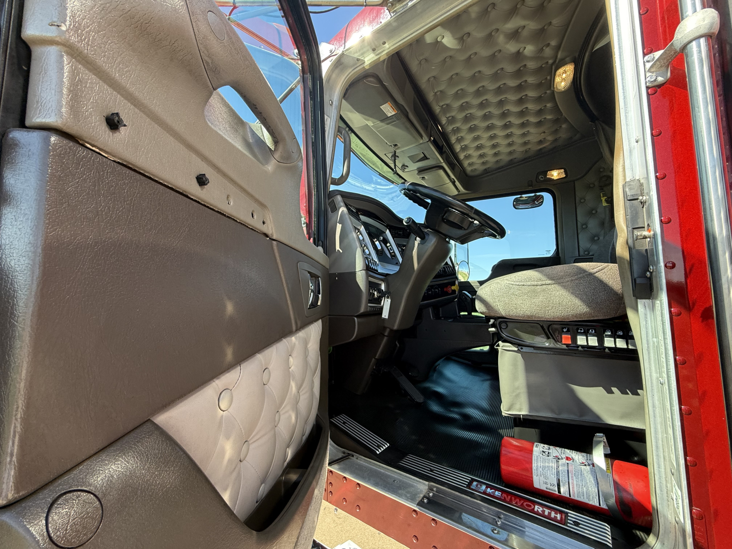 Kenworth W900L truck interior, driver seat and dashboard visible through open door; textured quilted upholstery on door panel, steering wheel, pedals, and seat; flame-red exterior frame visible
