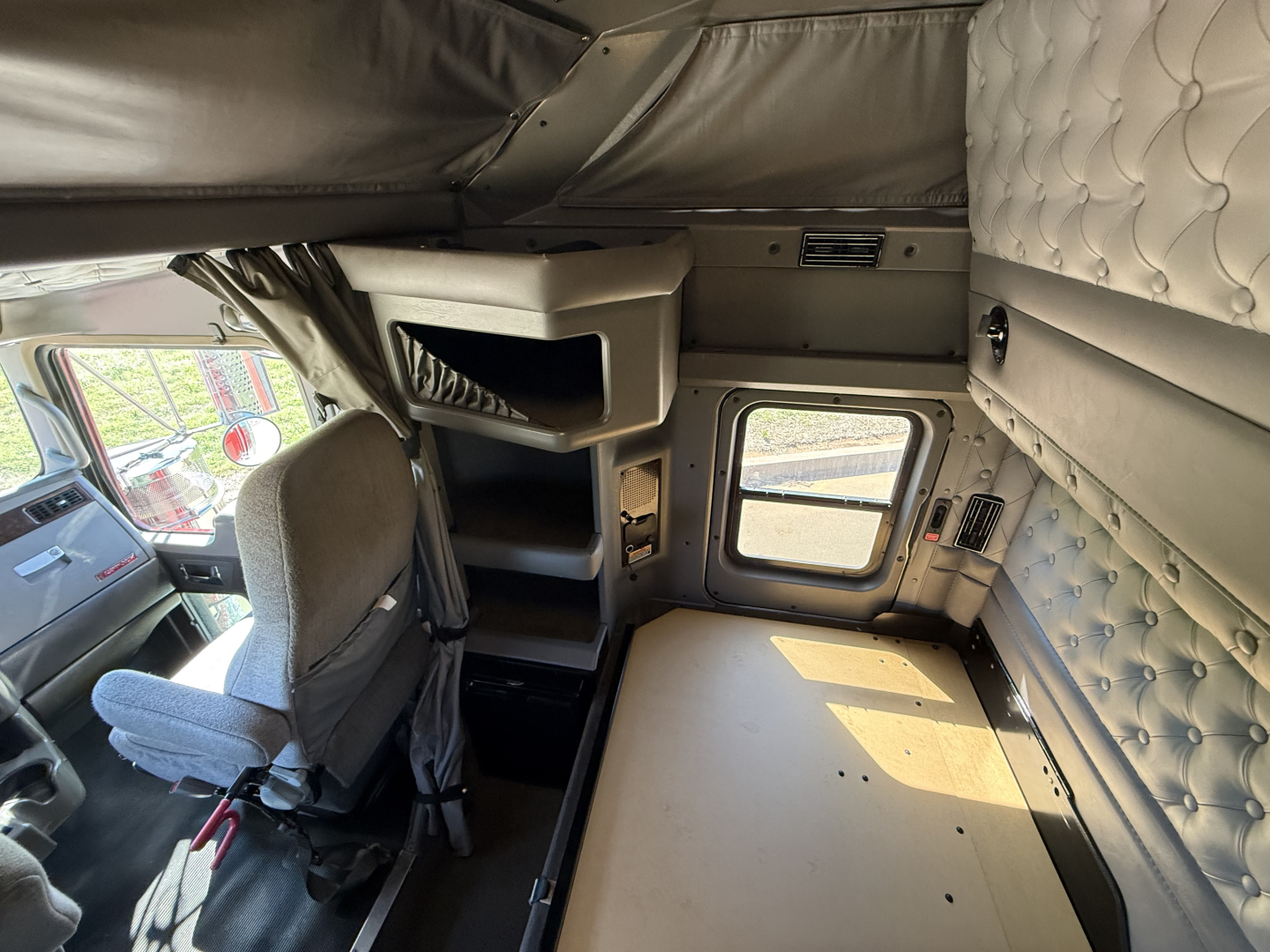 Kenworth W900L truck sleeper cab interior showing beige quilted side panels, overhead bunk area, storage shelves, and a small window; driver's seat and dashboard partially visible.