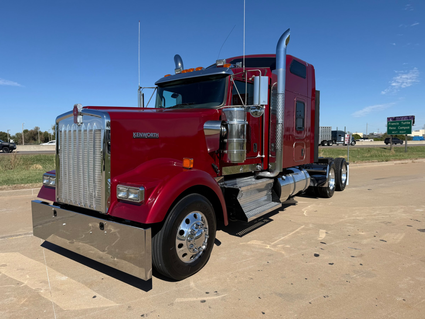 Kenworth W900L truck in red with chrome grille, dual exhaust stacks, and large front bumper. Visible text includes 'KENWORTH' on grille and side, with a chrome fuel tank and steps.