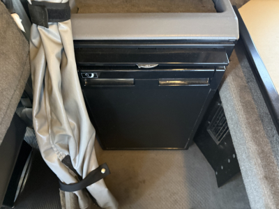 Kenworth W900L cab interior showing a black rectangular cabinet or mini-fridge under a sleeper area; surrounding upholstery and storage space.