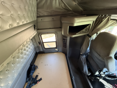 Kenworth W900L sleeper cab interior cargo area with quilted wall padding and passenger seat visible; small doorway and overhead storage; bare floor behind driver seat.