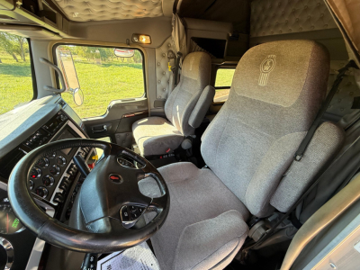 Truck interior of a Kenworth W900L showing steering wheel, driver's seat, and passenger seat with light gray fabric. Dashboard with gauges and controls visible on left.