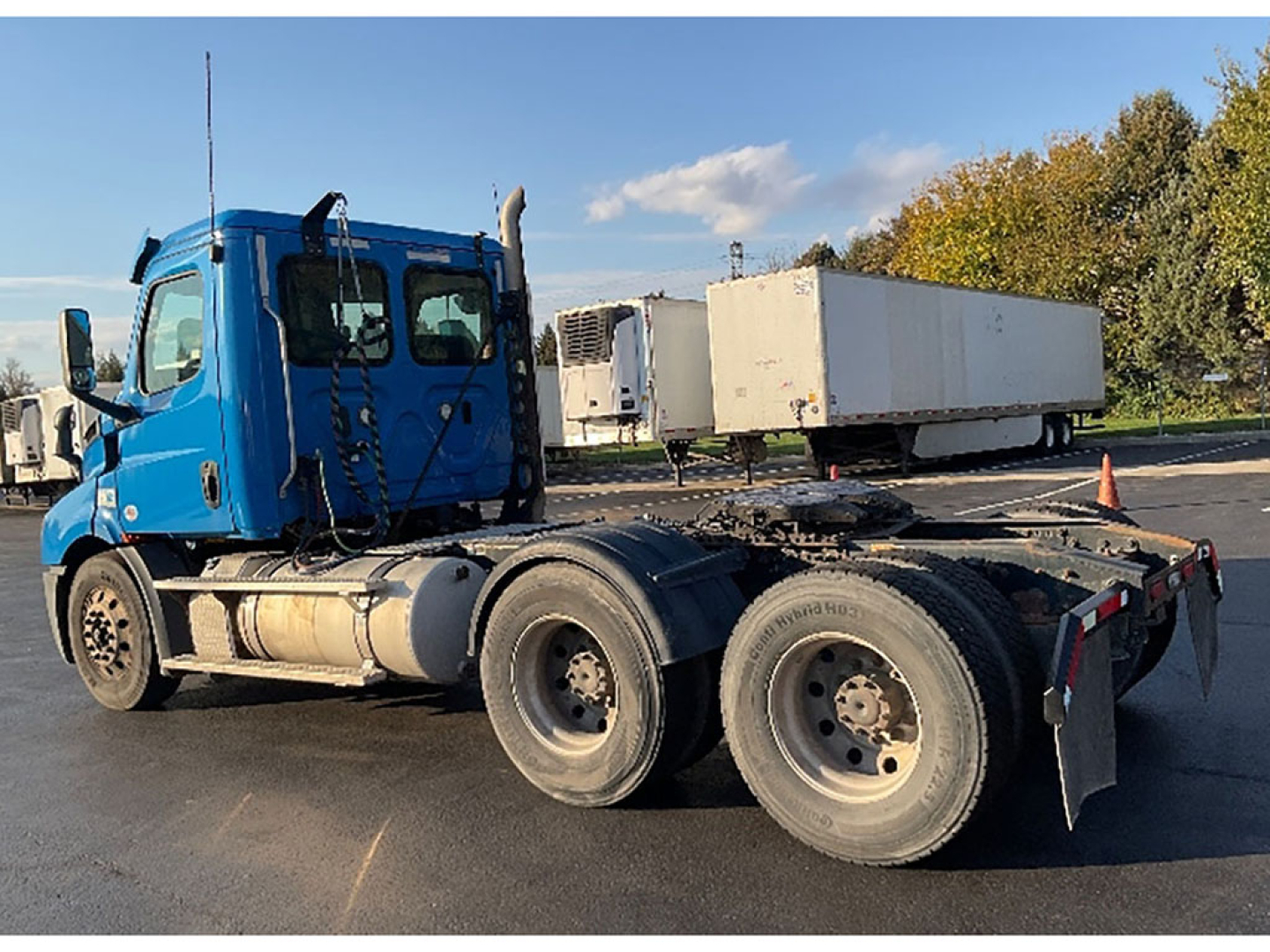 Blue Freightliner Cascadia 116 day cab tractor with dual rear axles, side view showing fuel tanks and wheel wells, attached box trailer in background.