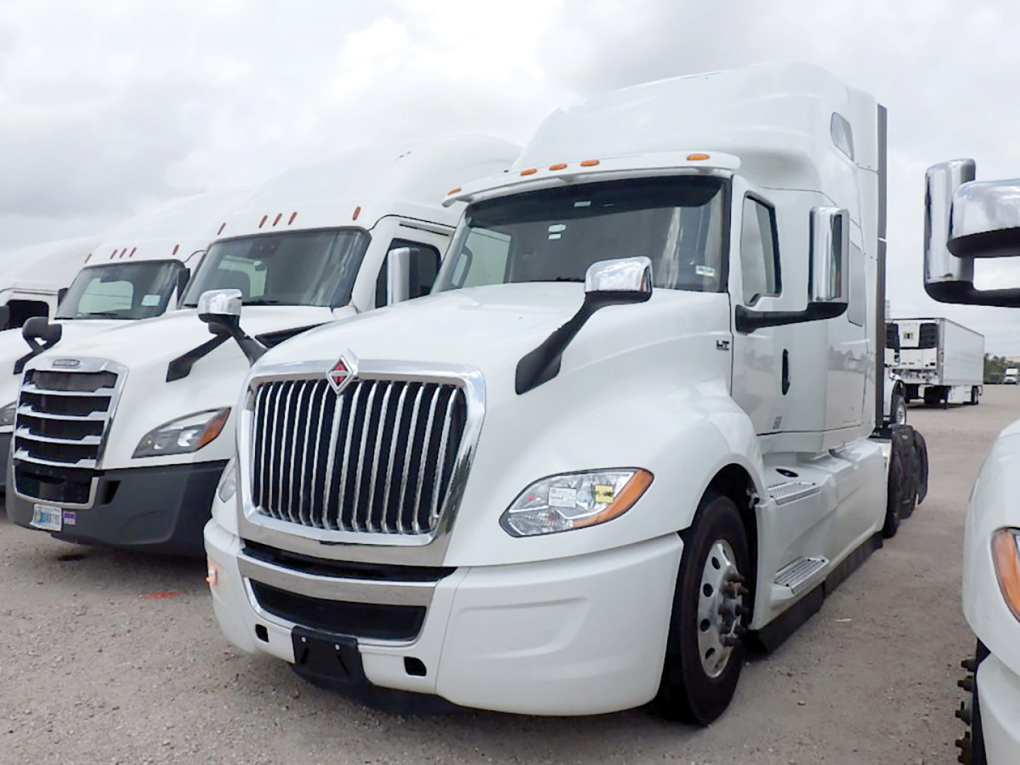 International LT625 conventional sleeper truck in a parking lot, white cab, chrome grille, side mirrors, front bumper, and other trucks visible in background.