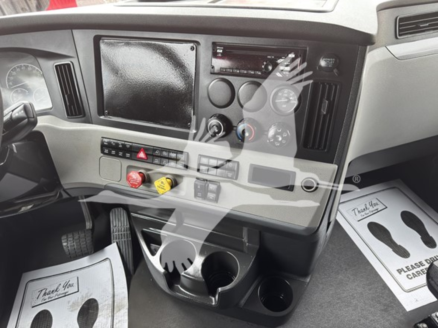 Dashboard of a Freightliner Cascadia 126 truck interior showing steering column, center console with controls, large display screen, various switches, cup holders, and paper mats on the floor.