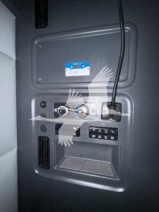 Cab interior side panel with control console and connectors; small blue warranty/ID sticker on upper panel; gauges and switches arranged below; vertical cable leading to a socket.