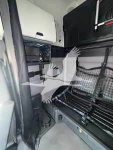 Cab interior of a Freightliner Cascadia sleeper showing bunk area with storage cabinet and mattress platform, side nets, and control panels.