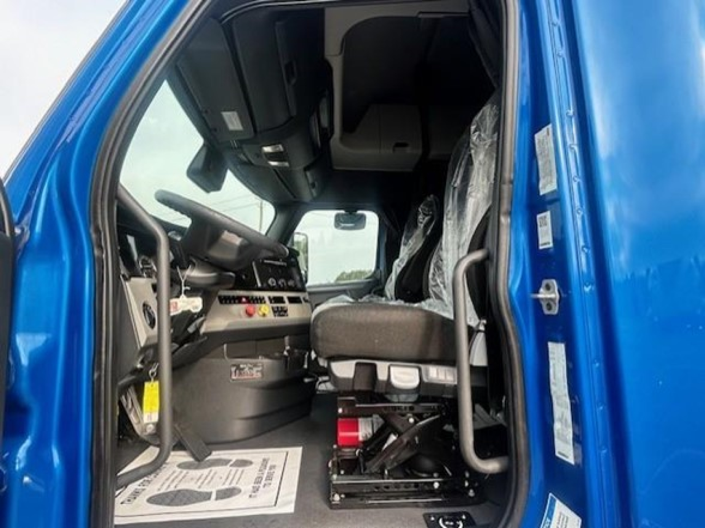 Truck cab interior of a blue 2022 Freightliner Cascadia 126 showing driver's seat, dashboard, and entry with plastic seat covers.