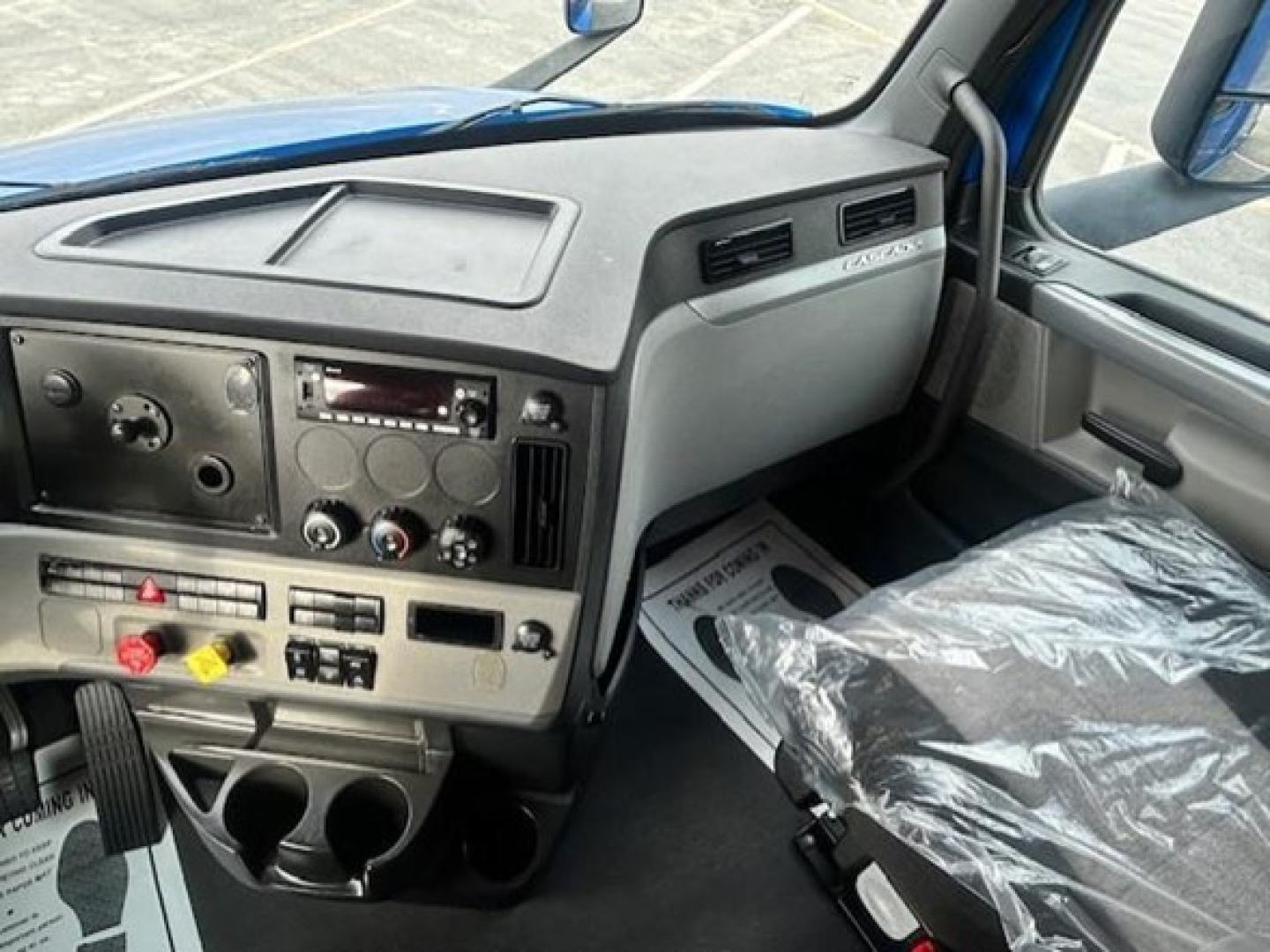 Interior of a Freightliner Cascadia truck cab showing dashboard controls, radio, climate knobs, cup holders, and plastic-covered seat.