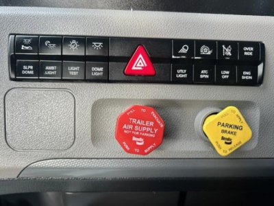 Dashboard control panel of a Freightliner truck showing various switches and indicators: red triangle hazard button, red engine air supply reservoir cap, yellow parking brake cap, and labeled switches including light, dome light, utility li