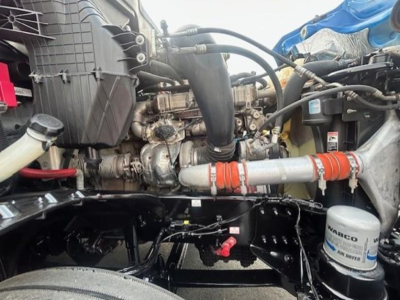 Engine bay of a Freightliner Cascadia showing intake, hoses, and a white coolant pipe with orange couplings; visible filter canister labeled MACKO or similar, various black metallic components and wiring.