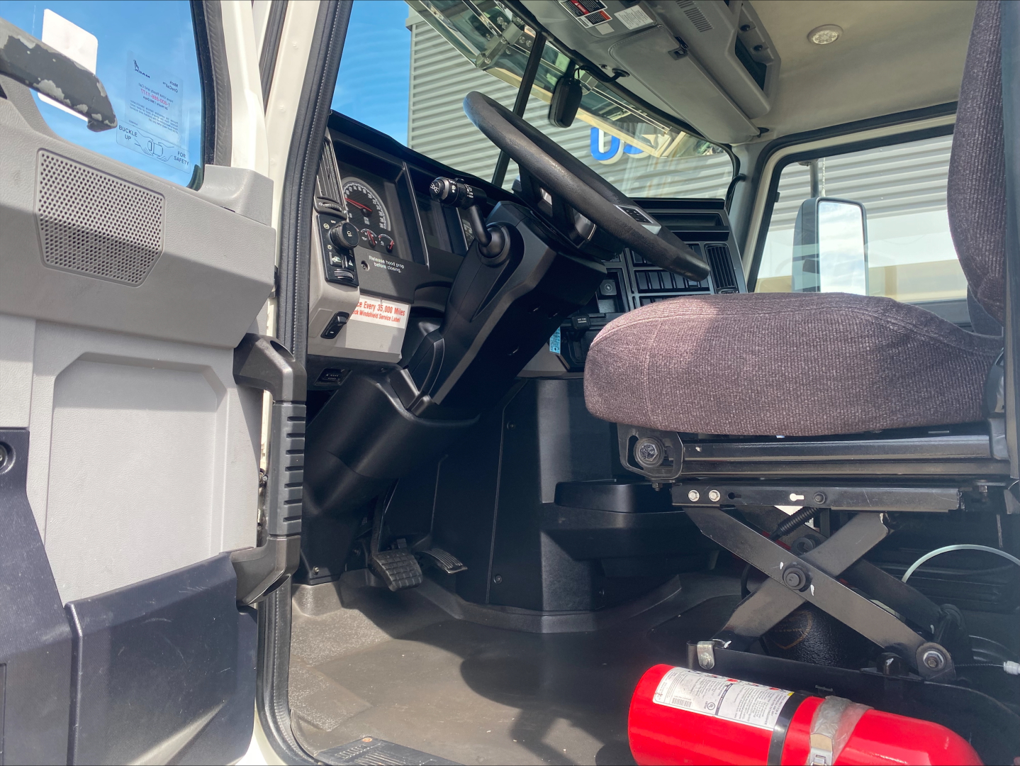 Truck cab interior of a 2021 Mack AN42T showing driver's seat, steering wheel, dashboard, and open door; visible pedals and fire extinguisher on the floor.