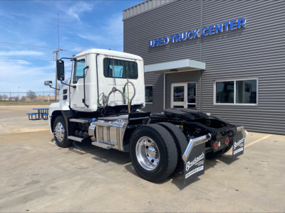 Truck chassis of a 2021 Mack AN42T daycab seen from rear-left, with dual rear axles, chrome fuel tanks, and wheel wells; building in background with USED TRUCK CENTER sign.