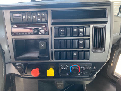 Dashboard of a 2021 Mack AN42T showing controls, switches, and climate dials. Visible text includes R N D P and Mack-branded gear selector, battery-like red and yellow knobs, and HVAC controls.