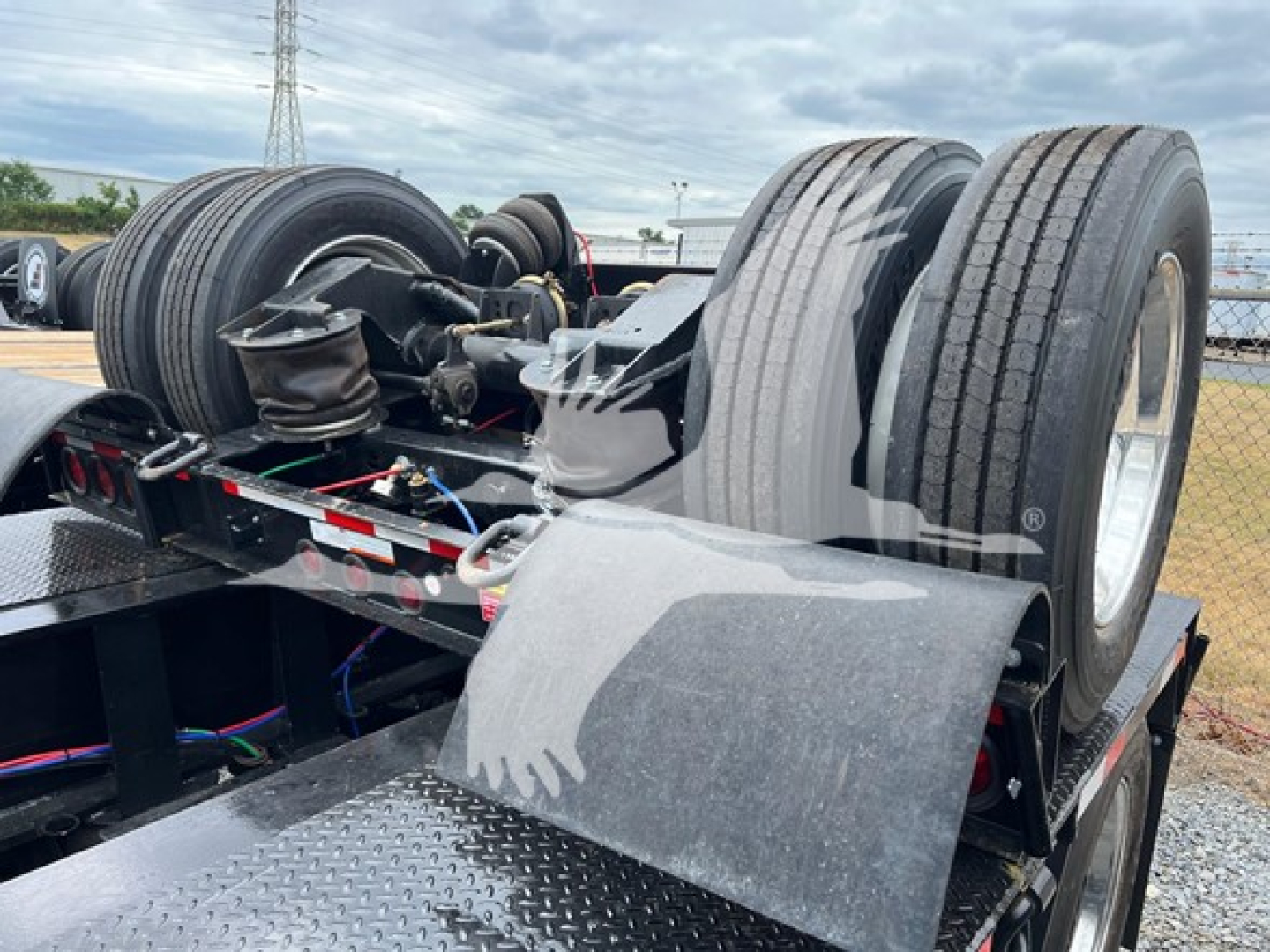 Lowboy trailer rear section showing two tandem axles and dual tires, hydraulic components, and fender; visible chain and hoses, textured aluminum deck, and wheel hubs.