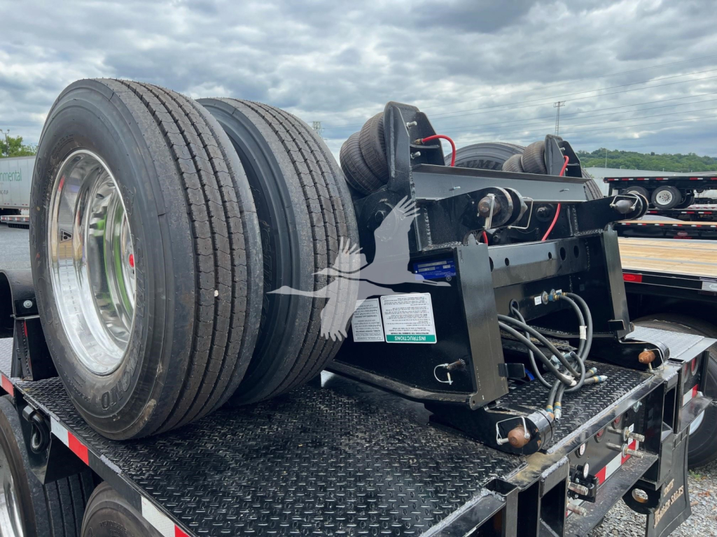 Trailer hitch mechanism on a lowboy trailer showing dual rear tires and mounting hardware with hoses and electrical connectors.