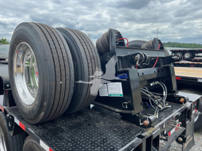 Trailer hitch mechanism on a lowboy trailer showing dual rear tires and mounting hardware with hoses and electrical connectors.