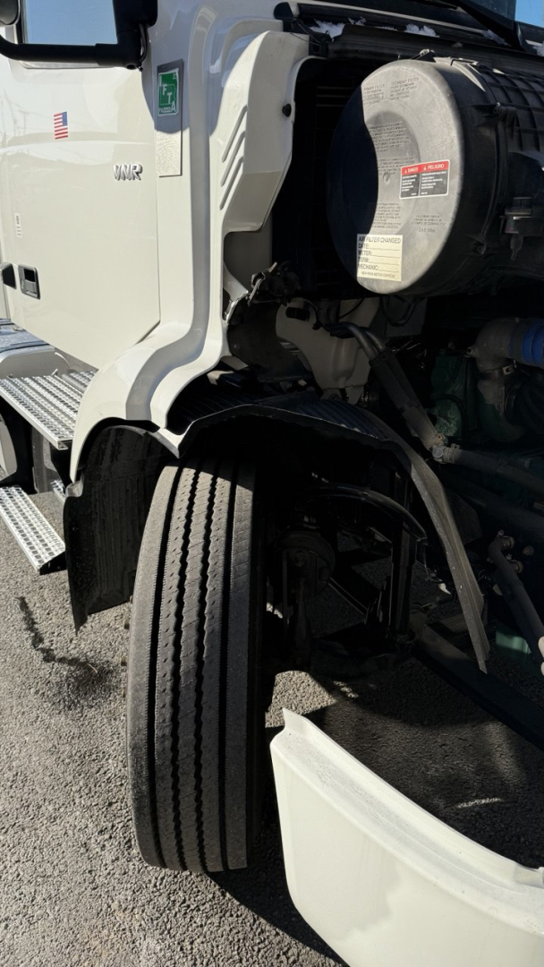 Front-left view of a Volvo VNR300 day cab truck showing front wheel well and diesel engine bay components with a circular air intake/def tank and warning labels.