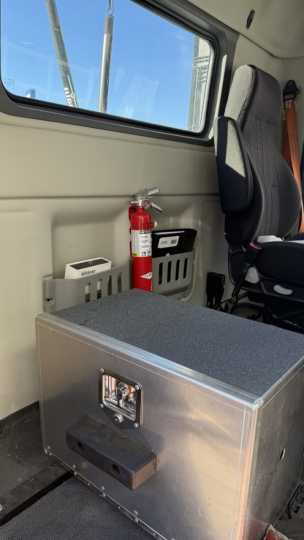 Vehicle interior showing a metal toolbox with latch, fire extinguisher, and seat in a Volvo VNR300 day cab unit compartment.