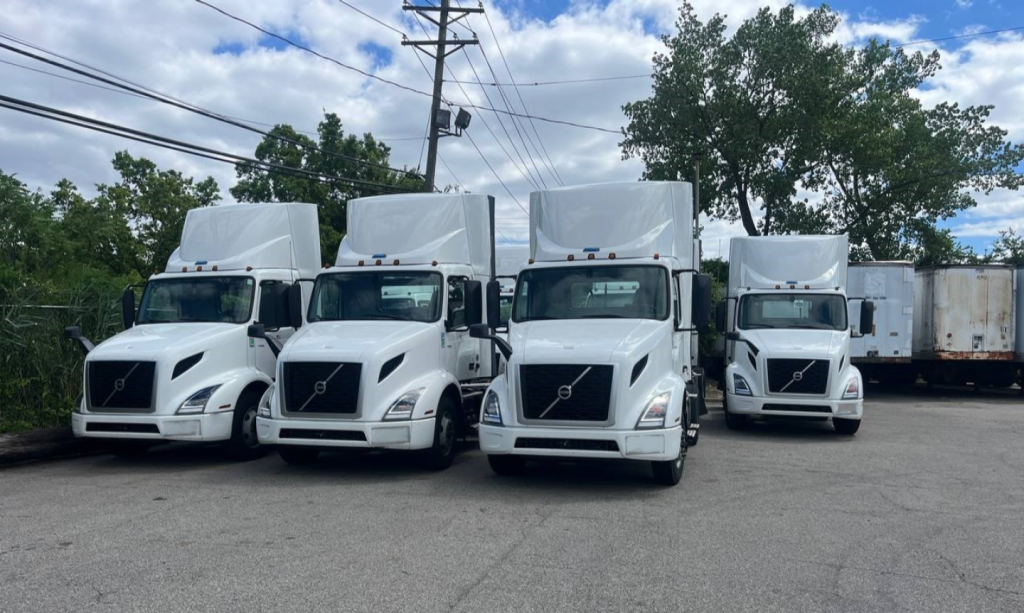 Front three-quarter view of a row of white Volvo VNR300 4x2 day cab trucks with cargo trailers in the background.