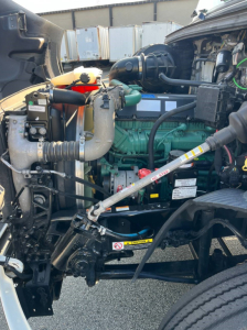 Volvo VNR300 engine bay showing green diesel engine, hoses, radiator, battery, and various fluid reservoirs in a daytime cab truck.