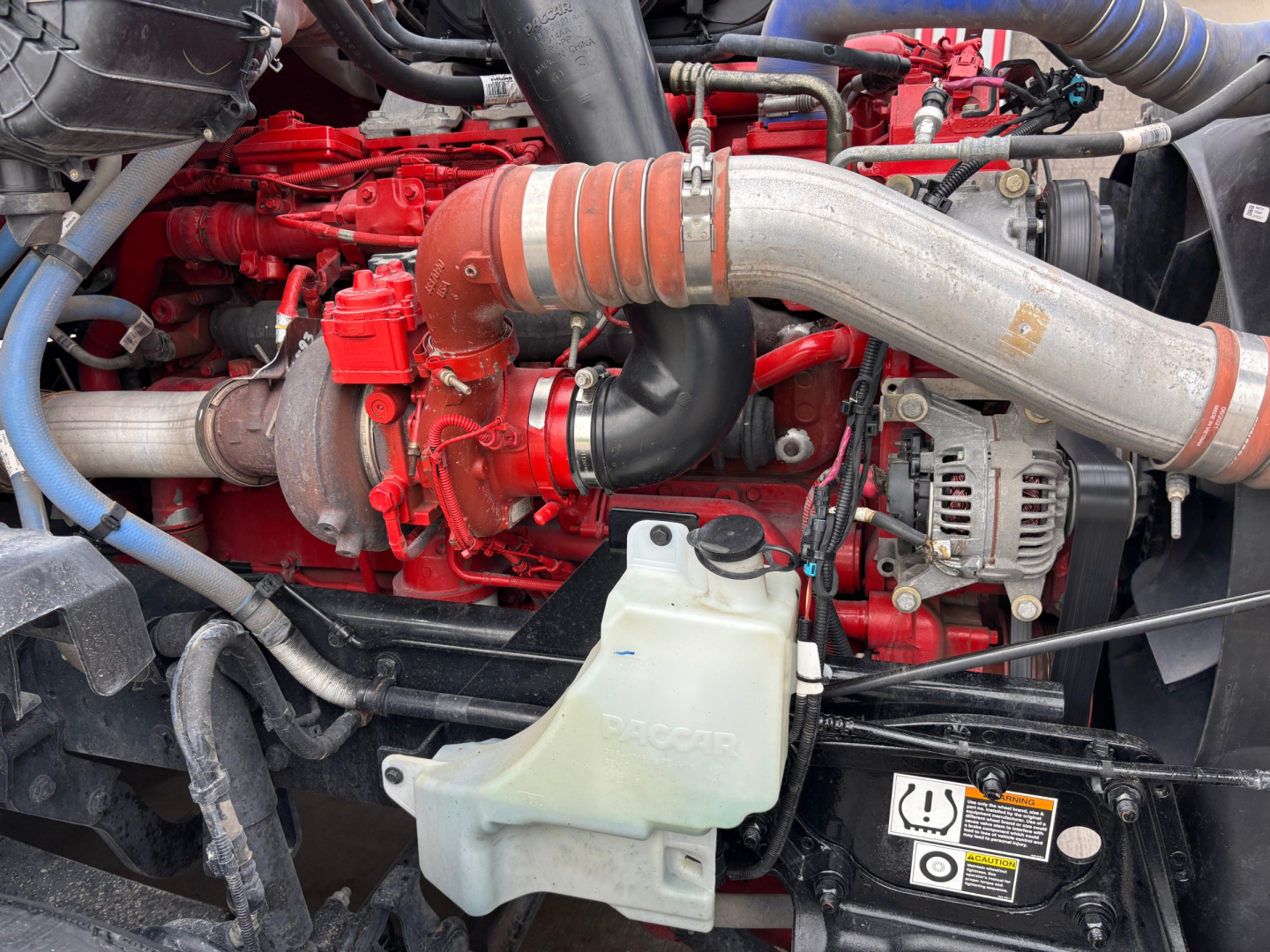 Kenworth T680 engine bay showing red engine block, large silver intake pipe, coolant reservoir, and alternator with wiring.