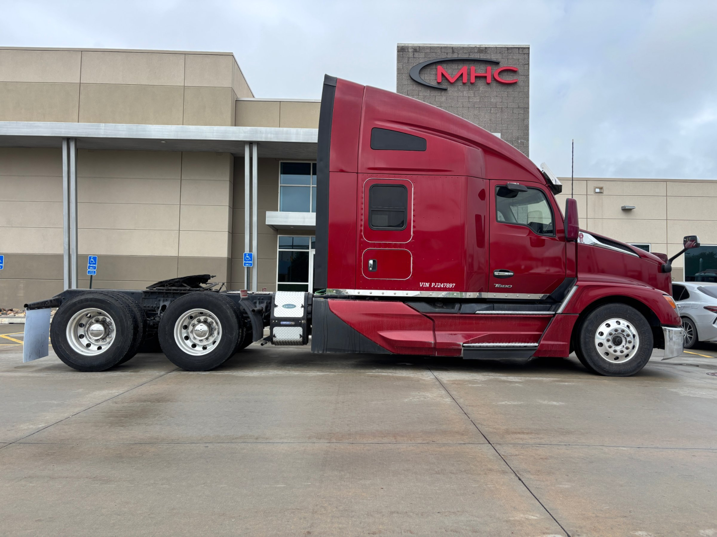 Kenworth T680 tractor in red side profile showing sleeper cab, tandem axle rear, and MHC dealership building in background.