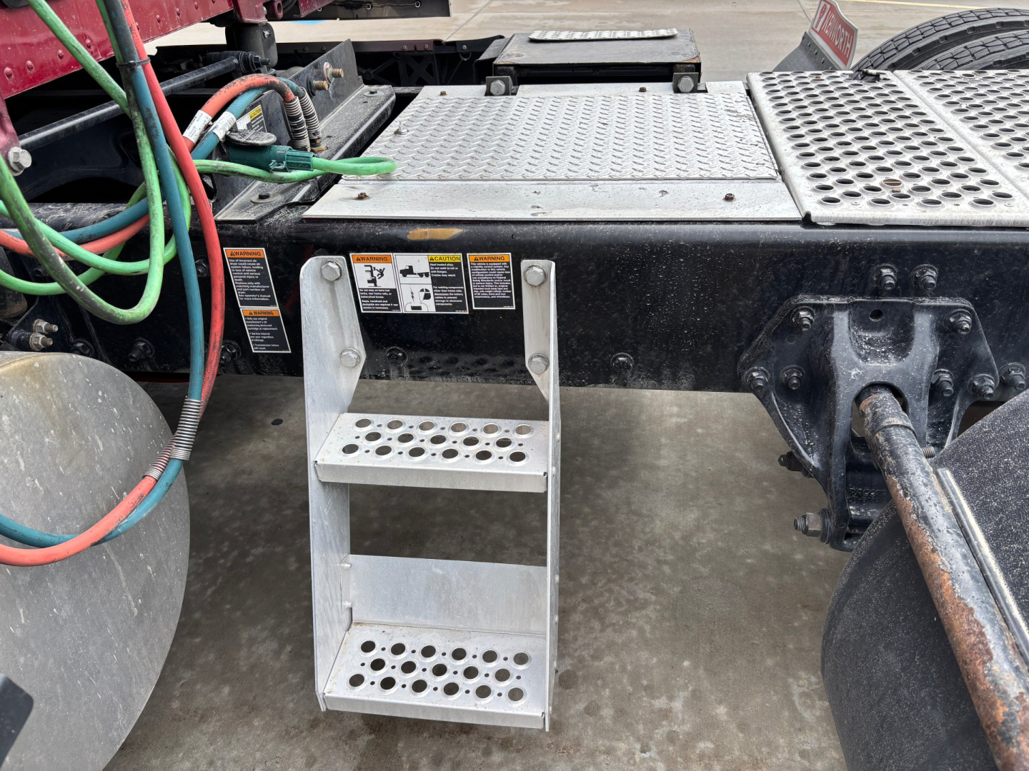 Two-step aluminum foothold attached to a Kenworth T680 truck chassis with perforated steps and warning labels, wiring harnesses, and a portion of the drive axle visible.